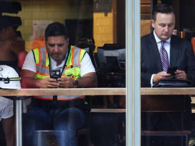 A man wearing high-visibility construction gear looks at his phone while seated next to a man in a business suit doing the same